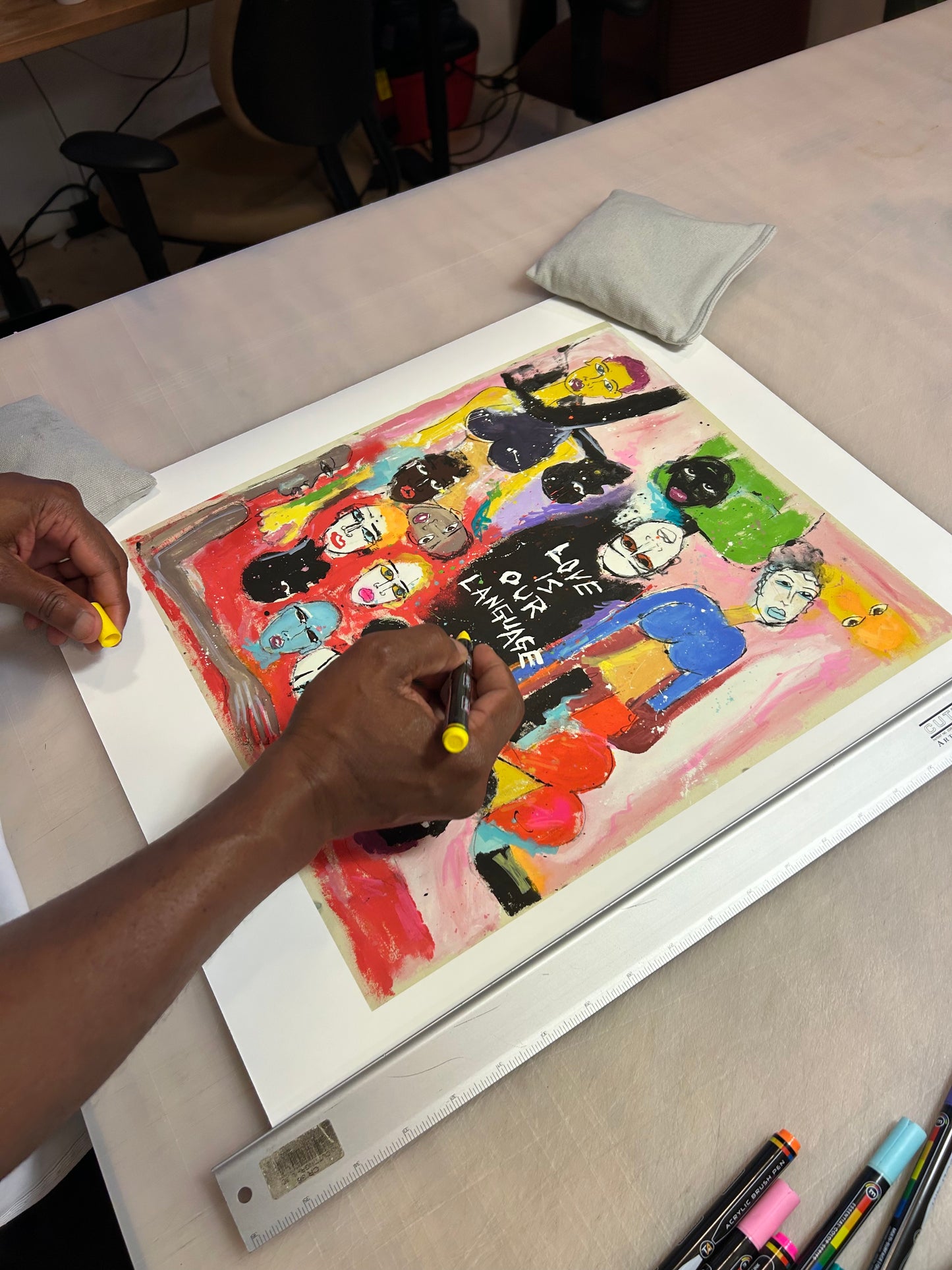Miles Regis' hand is drawing on his print, 'Love Is Our Language' at a work table.